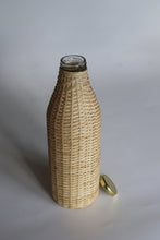 Load image into Gallery viewer, A clear glass bottle wrapped in a natural woven rattan or wicker sleeve, with a small gold-toned metal cap resting beside it on a light gray background.
