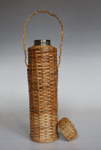 Load image into Gallery viewer, A tall cylindrical woven rattan or wicker bottle case with a carry handle and a separate matching lid placed beside it, photographed on a neutral gray studio background.
