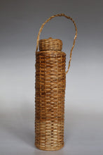 Load image into Gallery viewer, A tall, cylindrical woven rattan or wicker bottle case with a matching lid and a single loop handle, shown on a neutral gray studio background.
